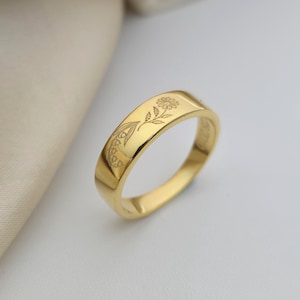 May include: A gold ring with engraved floral patterns. The ring has a flat band with a flower and leaf design. The ring is displayed on a white surface.