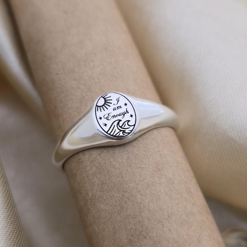 May include: A silver ring with a rounded oval face. The face features a circular design with a sun, waves, and the words "I am Enough." The ring is displayed on a neutral-toned surface.