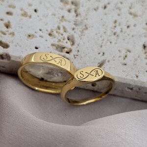 Pode incluir: Dois anéis de ouro, um em estilo de banda clássica e outro em estilo de sinete, são exibidos. Ambos os anéis apresentam as iniciais gravadas "SxA" com um símbolo de infinito estilizado. Eles estão em uma superfície clara e texturizada.