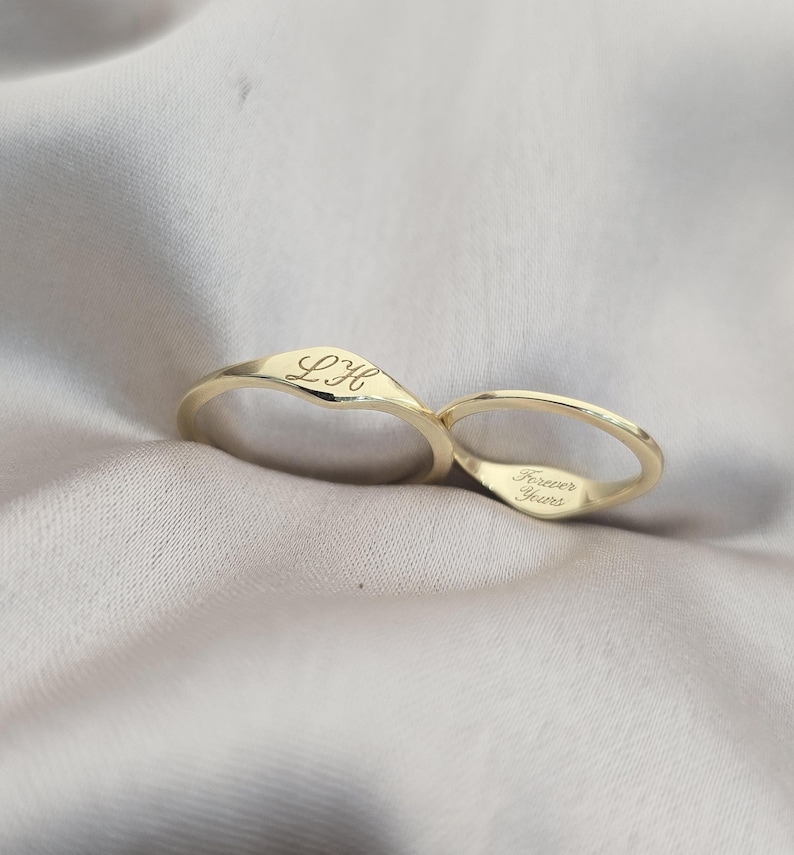 Op de afbeelding: Twee goudkleurige ringen met een gladde, gepolijste afwerking. E&eacute;n ring is voorzien van de gegraveerde initialen LH in cursief schrift op de buitenband, terwijl de andere ring de gegraveerde tekst Forever Yours aan de binnenzijde van de band toont.