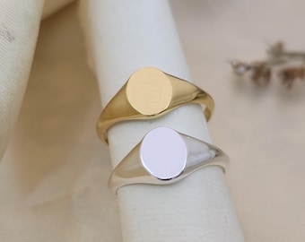 Könnte beinhalten: Zwei Siegelringe, einer goldfarben und einer silberfarben, werden gezeigt. Jeder Ring hat eine glatte, ovale Oberfläche und ein schlichtes Band. Die Ringe werden auf einer weißen Oberfläche mit einem weichen, neutralen Hintergrund präsentiert.