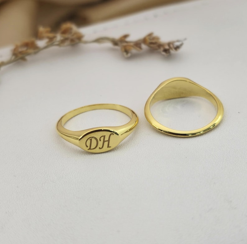 Puede incluir: Dos anillos de sello dorados. Un anillo tiene las iniciales "DH" grabadas en una cara ovalada. El otro anillo tiene una cara ovalada lisa. Ambos anillos est&aacute;n sobre una superficie blanca, con una ramita borrosa de flores secas en el fondo.