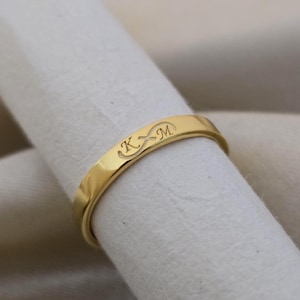May include: A gold ring with the engraved initials "K" and "M" and an infinity symbol. The ring is displayed on white fabric, highlighting its polished surface and simple design. The ring is a band style, suitable for everyday wear.