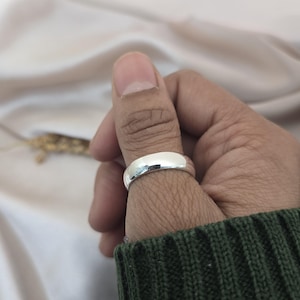 Puede incluir: Un anillo de plata pulida con una banda redondeada se lleva en un dedo. El anillo refleja la luz, creando una apariencia brillante y brillante. El fondo es de un color suave y neutro.