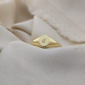 Puede incluir: Un anillo de sello dorado con una cara ovalada lisa. El anillo presenta una peque&ntilde;a "a" grabada en la cara. El anillo est&aacute; sobre un fondo de tela suave y blanquecina.