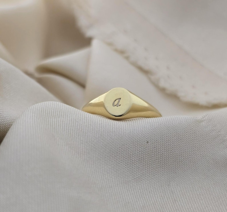 Puede incluir: Un anillo de sello dorado con una cara ovalada lisa. El anillo presenta una peque&ntilde;a "a" grabada en la cara. El anillo est&aacute; sobre un fondo de tela suave y blanquecina.