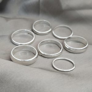Puede incluir: Siete anillos de plata de diferentes anchos se muestran sobre una tela gris. Los anillos son circulares con una superficie pulida y reflectante. Los anillos probablemente est&aacute;n hechos de plata de ley y son adecuados para el uso diario.