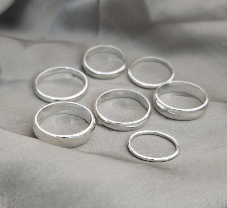 Puede incluir: Siete anillos de plata de diferentes anchos se muestran sobre una tela gris. Los anillos son circulares con una superficie pulida y reflectante. Los anillos probablemente est&aacute;n hechos de plata de ley y son adecuados para el uso diario.