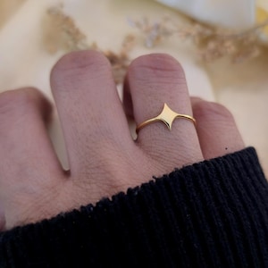 May include: A gold ring featuring a geometric starburst design. The ring is worn on a finger, highlighting its slender band and the symmetrical shape of the starburst. The ring is a piece of jewellery.