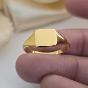 May include: A gold signet ring with a square face. The ring has a polished finish and a simple, classic design. The ring is likely made of gold and is a piece of jewelry.