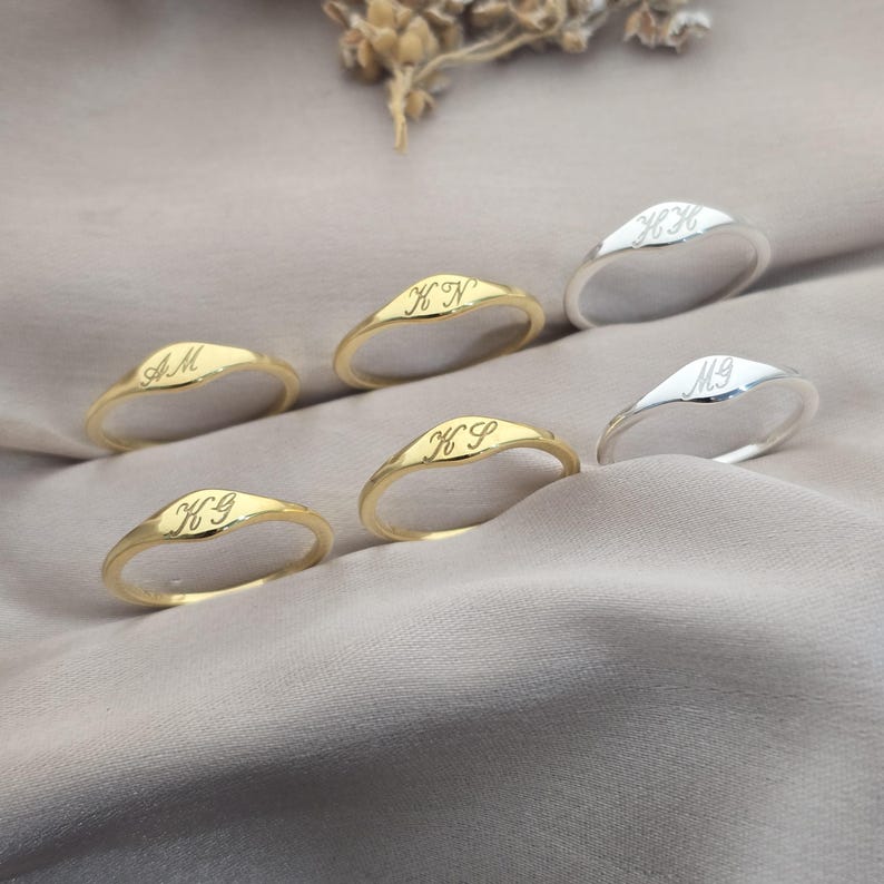 Op de afbeelding: Een collectie van vijf dunne metalen ringen, drie in goudkleur en twee in zilverkleur. Elke ring heeft een puntig, verhoogd middengedeelte gegraveerd met twee cursieve hoofdletters, waaronder AM, KN, KS, HH en MG, gerangschikt op gedrapeerde stof.