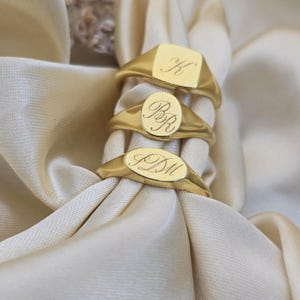 May include: Three gold signet rings with engraved initials. The top ring has a square face with the letter "K". The middle ring has an oval face with "BR". The bottom ring has an oval face with "PDM". The rings are displayed on a cream-colored fabric.