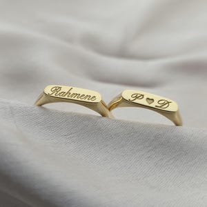 May include: Two gold-colored signet rings. One ring has the name "Rahmene" engraved on it. The other ring has the initials "P D" with a heart symbol engraved on it. The rings are displayed on a white fabric.
