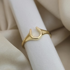 Op de afbeelding: Een goudkleurige ring met een hoefijzerontwerp. De ring is geplaatst tegen een witte en crèmekleurige achtergrond. Het hoefijzer is een traditioneel symbool van geluk.