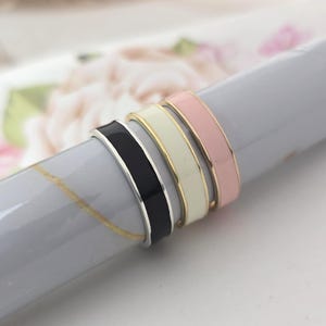 May include: Four enamel rings in black, cream, gold, and pink are stacked together. The rings have a flat band design with a shiny finish. The rings are displayed on a grey surface with a floral background.