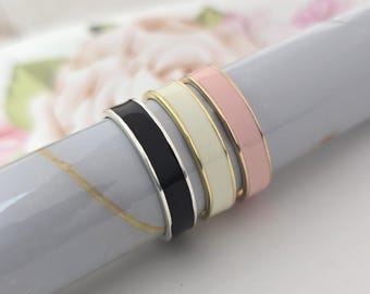 Puede incluir: Cuatro anillos de esmalte en negro, crema, dorado y rosa están apilados. Los anillos tienen un diseño de banda plana con un acabado brillante. Los anillos se muestran sobre una superficie gris con un fondo floral.