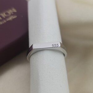 Puede incluir: Un anillo plateado con una parte superior rectangular y el n&uacute;mero "555" grabado. El anillo se muestra sobre una superficie blanca, con un vistazo a una caja granate en el fondo.