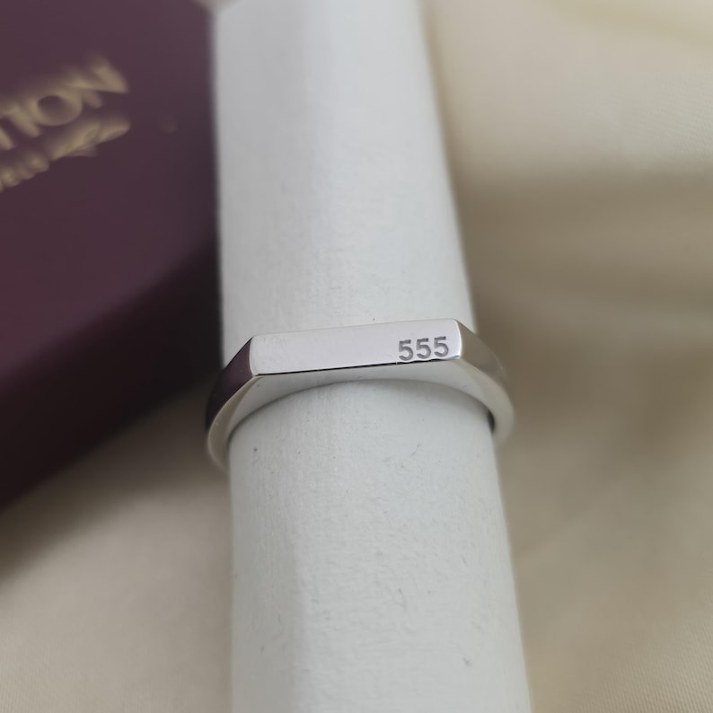 Puede incluir: Un anillo plateado con una parte superior rectangular y el n&uacute;mero "555" grabado. El anillo se muestra sobre una superficie blanca, con un vistazo a una caja granate en el fondo.