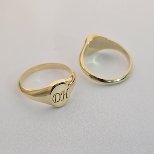 Puede incluir: Dos anillos de sello dorados. Un anillo tiene las iniciales grabadas "DH" en su cara ovalada. El otro anillo es liso. Ambos anillos son de oro amarillo brillante y est&aacute;n sobre un fondo neutro.