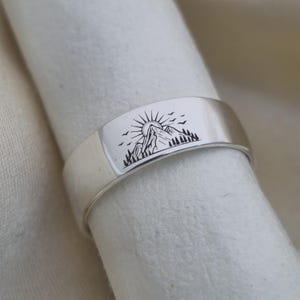 Puede incluir: Un anillo plateado con un diseño de montaña y sol. El anillo presenta una banda plana con una escena de paisaje grabada de una montaña, sol, pájaros y árboles. El anillo se muestra sobre una superficie blanca.