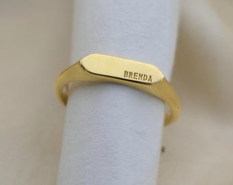 May include: A gold-colored signet ring with the name "BRENDA" engraved on the rectangular face. The ring is displayed on a white surface, showcasing its polished finish and simple design.