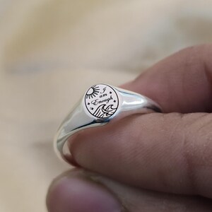 May include: A silver signet ring with a circular face. The face features a sun, waves, and stars, along with the words "I am Enough". The ring has a polished finish and a smooth band.