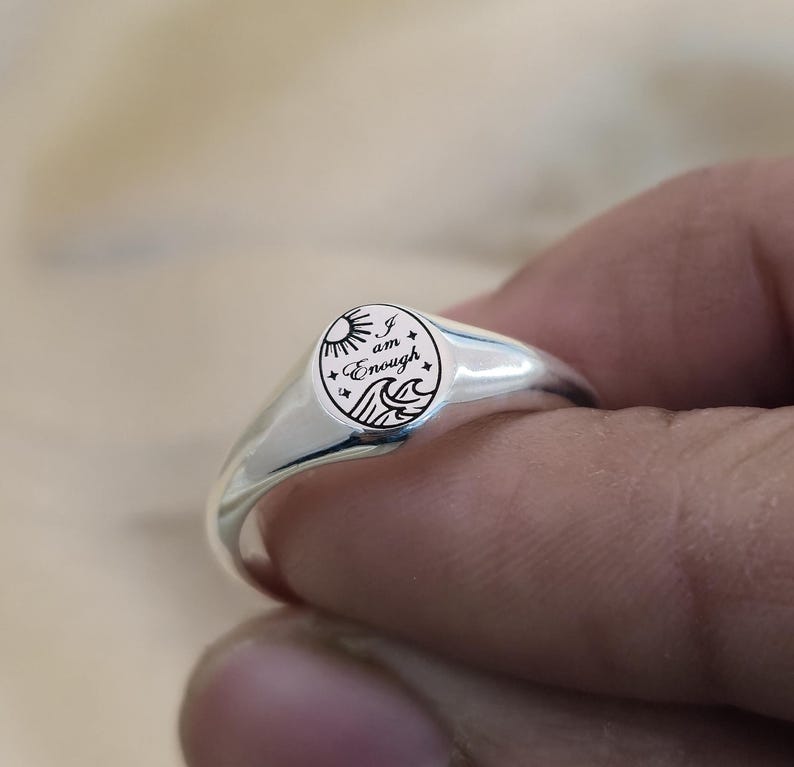 May include: A silver signet ring with a circular face. The face features a sun, waves, and stars, along with the words "I am Enough". The ring has a polished finish and a smooth band.