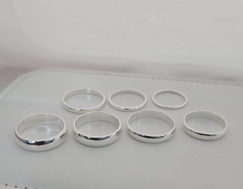 Puede incluir: Una colecci&oacute;n de siete anillos de plata de diferentes tama&ntilde;os, dispuestos sobre una superficie gris claro. Los anillos tienen un acabado liso y pulido, que refleja la luz ambiental. Los anillos probablemente son de plata de ley o un metal similar.