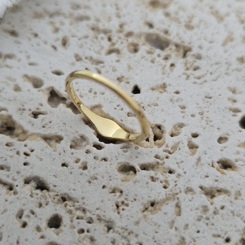 Op de afbeelding: Een gepolijste goudkleurige ring met een gladde, slanke band en een plat, langwerpig ovaalvormig oppervlak. De ring heeft een reflecterende metalen afwerking en een eenvoudig, doorlopend ontwerp dat over de gehele omtrek een constante breedte behoudt.