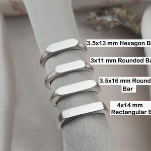 Könnte beinhalten: Vier silberne Ringe mit rechteckigen und abgerundeten Stegen werden gezeigt. Die Ringe haben unterschiedliche Größen, mit Text, der Abmessungen wie 3,5x13 mm Hexagon Bar und 4x14 mm Rectangular Bar angibt. Die Ringe liegen auf einem grauen Stoff.