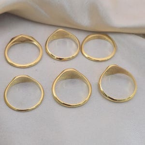 Puede incluir: Seis anillos de sello dorados. Cada anillo tiene una banda lisa y redondeada y una cara plana de forma ovalada. Los anillos se muestran sobre una superficie clara y texturizada.