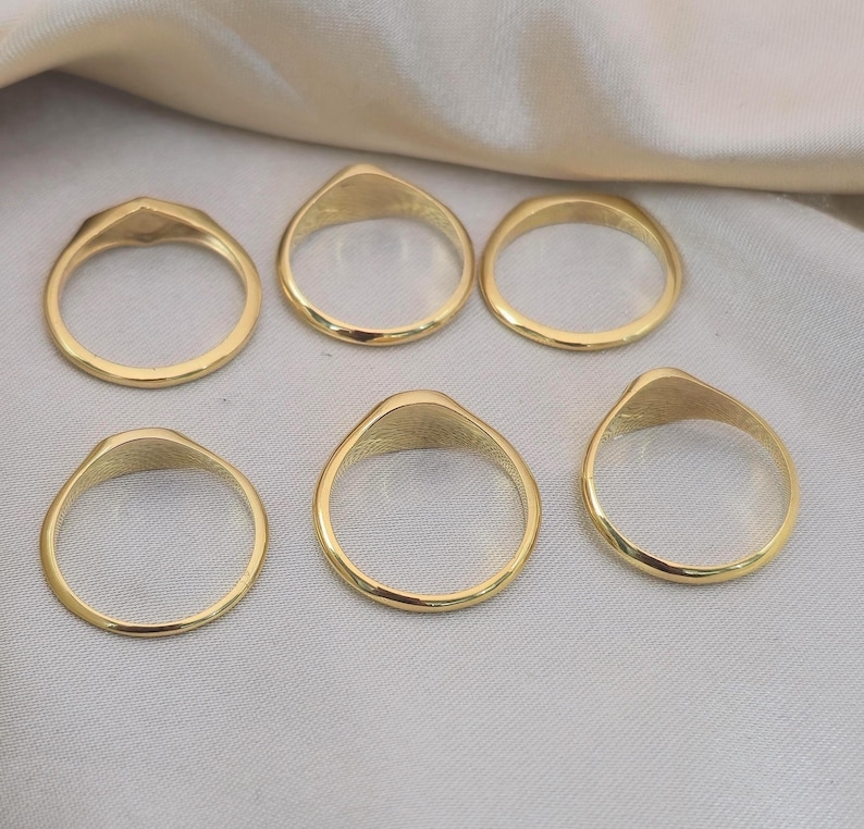 Puede incluir: Seis anillos de sello dorados. Cada anillo tiene una banda lisa y redondeada y una cara plana de forma ovalada. Los anillos se muestran sobre una superficie clara y texturizada.