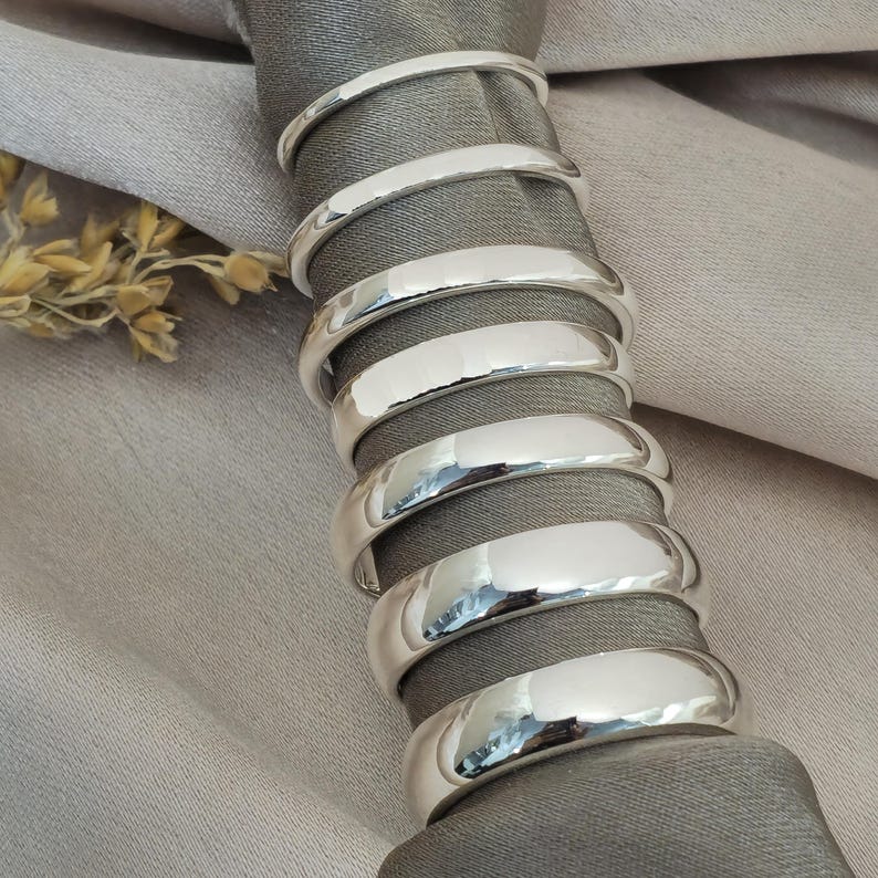 Puede incluir: Una pila de ocho servilleteros plateados brillantes. Los anillos son lisos y reflectantes, con una textura martillada en la superficie exterior. Est&aacute;n dispuestos alrededor de una servilleta gris, con un fondo beige.