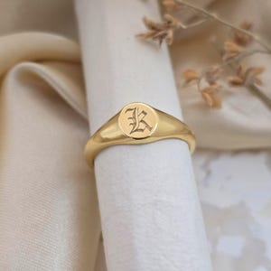 Könnte beinhalten: Ein goldener Siegelring mit poliertem Finish. Der Ring hat eine kreisförmige Oberfläche mit dem Buchstaben "K" in einer eleganten, stilisierten Schriftart. Der Ring wird auf einer weißen Oberfläche mit einem weichen, neutralen Hintergrund präsentiert.