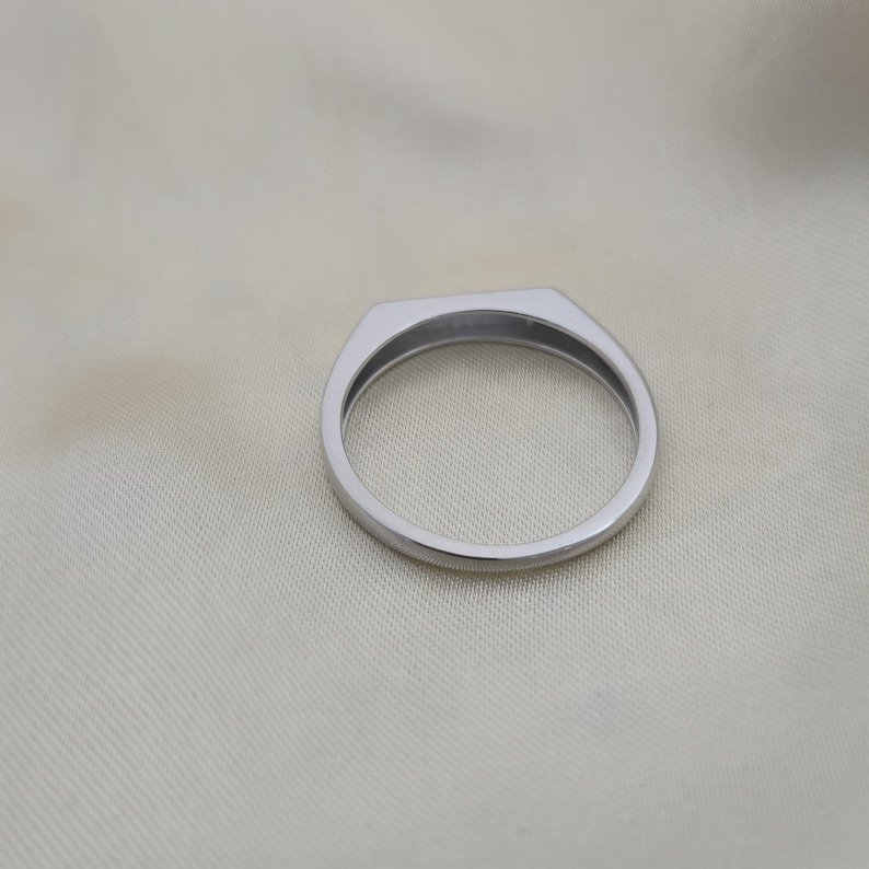 Puede incluir: Un anillo plateado con una parte superior plana y rectangular y una banda lisa y redondeada. El anillo est&aacute; hecho de un material met&aacute;lico brillante y se coloca sobre un fondo blanco roto.