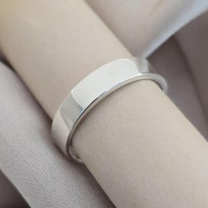 Peut inclure: Une bague en argent poli avec un dessus plat et rectangulaire. La bague est présentée sur un tissu beige, soulignant sa surface réfléchissante. L'anneau est lisse et simple, mettant en valeur un design minimaliste. Ce bijou est adapté au quotidien.