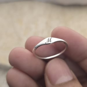 Könnte beinhalten: Ein silberner Ring mit glatter, polierter Oberfläche. Der Ring hat eine leicht gebogene rechteckige Oberfläche mit dem Buchstaben "M" in zarter Schrift eingraviert. Der Ring wird in einer Hand gehalten.
