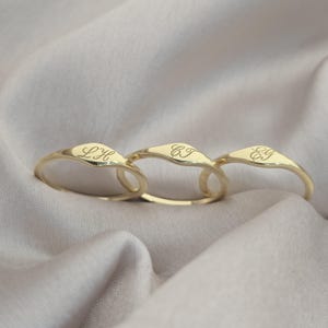 Op de afbeelding: Drie goudkleurige zegelringen met gladde, gepolijste oppervlakken. Elke ring is gegraveerd met een unieke set van twee cursieve initialen: LH, CJ en EG. De ringen hebben een afgeronde band en een licht verhoogd, plat vlak voor de letters.