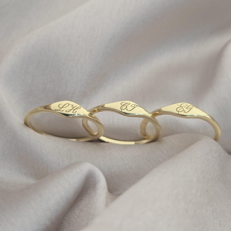 Op de afbeelding: Drie goudkleurige zegelringen met gladde, gepolijste oppervlakken. Elke ring is gegraveerd met een unieke set van twee cursieve initialen: LH, CJ en EG. De ringen hebben een afgeronde band en een licht verhoogd, plat vlak voor de letters.