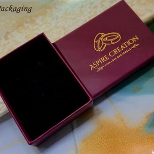 Puede incluir: Una caja de joyer&iacute;a burdeos abierta con un interior de terciopelo negro. La tapa de la caja presenta letras doradas y las palabras "Aspire Creation, Your Love Our Jewels" y dos anillos dorados. La palabra "Packaging" est&aacute; en la esquina superior izquierda.