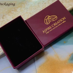 May include: An open, burgundy jewelry box with a black velvet interior. The box lid features the gold text "Aspire Creation" and two gold rings. The words "Your love our jewels" are below the logo.