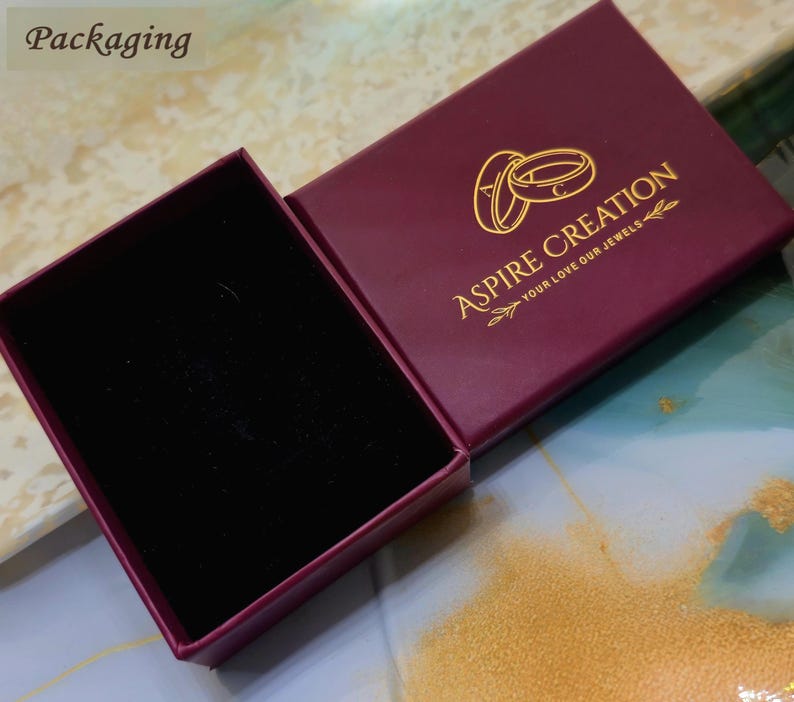 May include: An open, burgundy jewelry box with a black velvet interior. The box lid features the gold text "Aspire Creation" and two gold rings. The words "Your love our jewels" are below the logo.