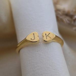 Puede incluir: Un anillo dorado con un diseño dividido. Cada extremo del anillo presenta un diseño en forma de corazón con las letras "J" y "K" grabadas. El anillo se muestra sobre una superficie blanca.