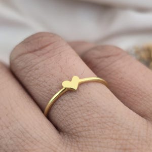 May include: A gold ring with a small heart-shaped charm. The ring has a slender band and a polished finish, creating a simple yet elegant design. The heart charm is centered on the ring, adding a touch of romance.