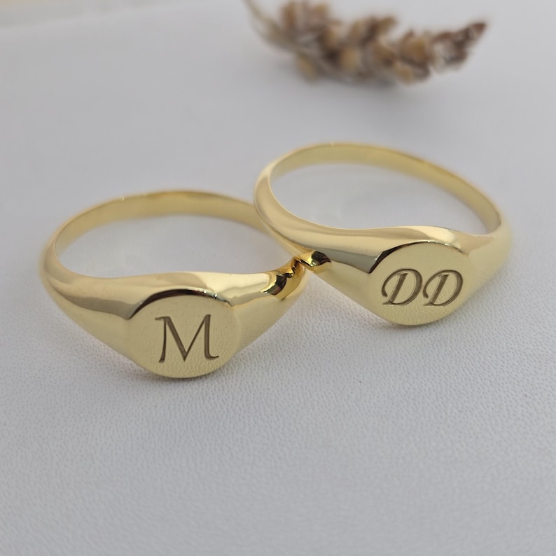 Puede incluir: Dos anillos de sello dorados. Un anillo tiene la letra "M" grabada en la cara ovalada, y el otro anillo tiene las iniciales "DD". Los anillos est&aacute;n sobre una superficie blanca.