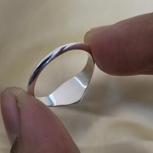 May include: A polished silver ring with a smooth, rounded band and a flat, rectangular face. The ring is held between two fingers, showcasing its reflective surface. The ring's design is simple and elegant.