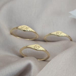 Op de afbeelding: Drie gouden ringen met een gladde, gepolijste afwerking en een V-vormig bovengedeelte. Elke ring is voorzien van een uniek gegraveerd monogram in cursief schrift: LH, CJ en EG. De ringen zijn dun en rond, met een consistente goudkleur.