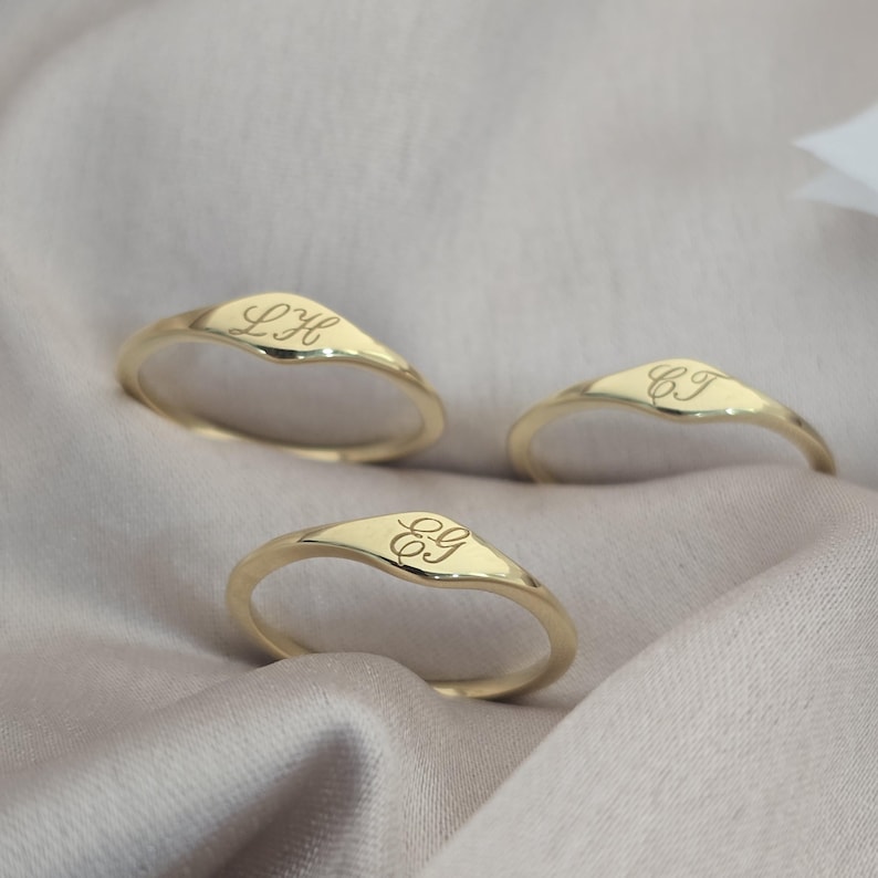Op de afbeelding: Drie gouden ringen met een gladde, gepolijste afwerking en een V-vormig bovengedeelte. Elke ring is voorzien van een uniek gegraveerd monogram in cursief schrift: LH, CJ en EG. De ringen zijn dun en rond, met een consistente goudkleur.