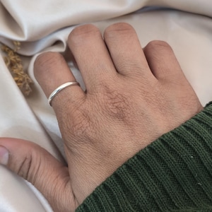 Puede incluir: Un anillo de plata liso se lleva en un dedo. El anillo es una banda sencilla. La persona lleva un jersey verde. El fondo es una tela beige claro.