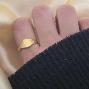 May include: A gold signet ring with a rectangular face and two small, sparkling stones. The ring is a classic design, suitable for everyday wear. The ring is shown on a finger.